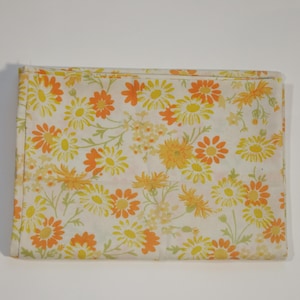 Puede incluir: Tela rectangular doblada con un estampado floral. El diseño presenta margaritas amarillas y naranjas con tallos y hojas verdes sobre un fondo blanco. La tela es probablemente de algodón o un material similar.