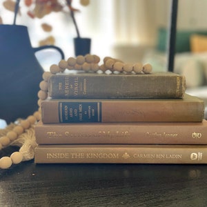 May include: A stack of four books with brown and beige covers. The top book has the title "The Aeneid of Virgil" on the spine. The second book has the title "Modern Arms and Free Men" on the spine. The third book has the title "The Secrets of My Life" on the spine. The bottom book has the title "Inside the Kingdom" on the spine. A wooden bead garland is in the foreground.