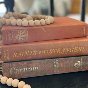 Puede incluir: Una pila de tres libros antiguos de tapa dura con cubiertas marrones y rojas. El libro superior tiene el título "The Innfolk" y el autor "Earls Buck". El libro del medio tiene el título "Saints and Strangers" y el autor "George F. Willison". El libro inferior tiene el título "Caravans" y el autor "James A. Michener".