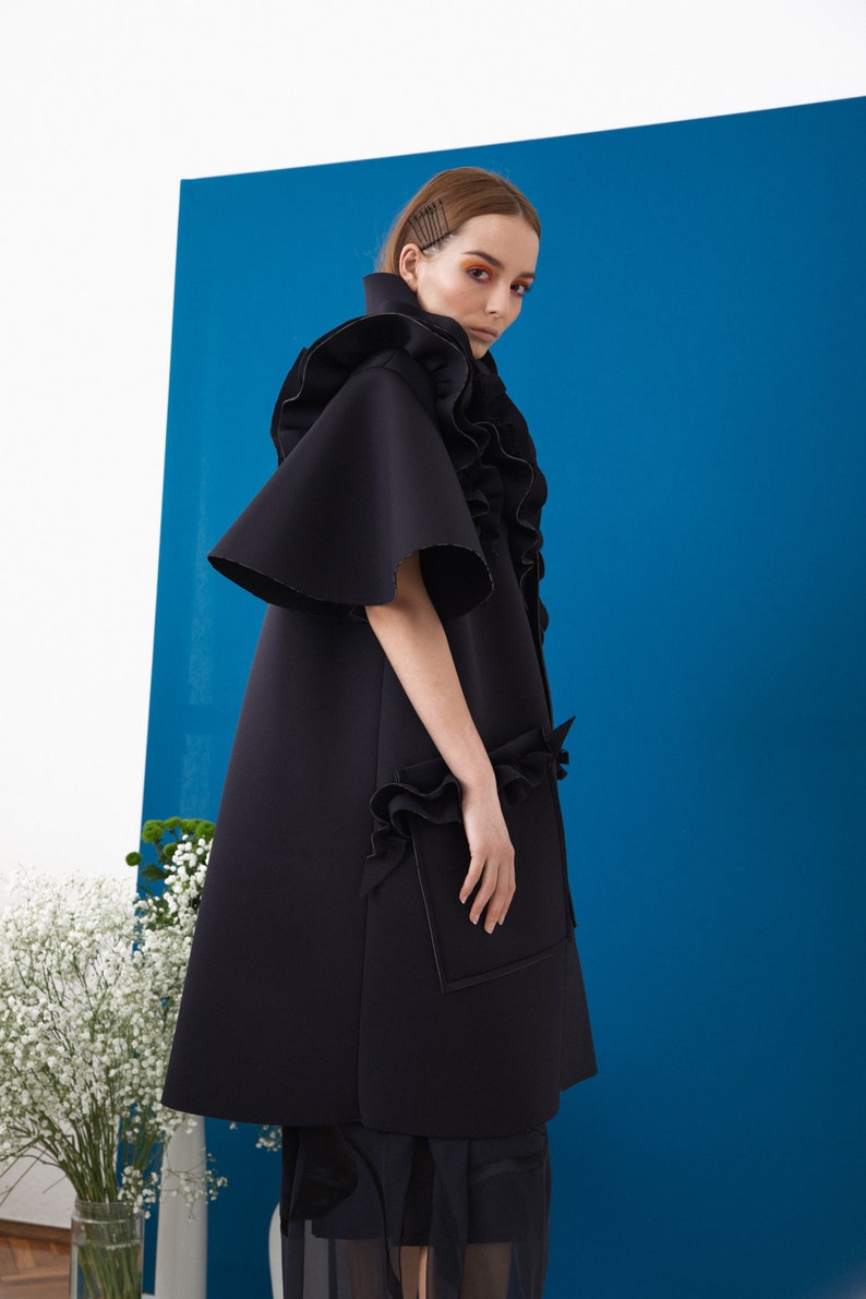 Pode incluir: Um casaco preto, com corte evas&ecirc;, com detalhes de babados e mangas curtas. O casaco apresenta uma gola alta e um design &uacute;nico, com um bolso na frente. O fundo &eacute; azul vibrante, com flores brancas num vaso.