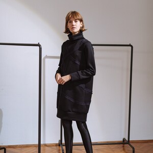 May include: A black turtleneck dress with a layered design and a ruffled hem. The dress is worn with black tights and black heeled shoes.