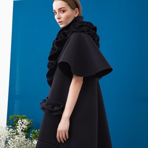 Pode incluir: Um casaco preto estilo capa com uma gola e mangas com babados dram&aacute;ticos. O casaco apresenta uma silhueta flare e um bolso com um detalhe de babados. O fundo &eacute; azul vibrante, com flores brancas no canto inferior esquerdo.