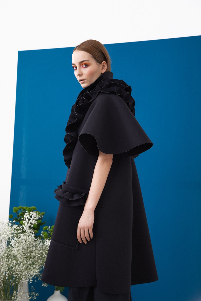Pode incluir: Um casaco preto estilo capa com uma gola e mangas com babados dram&aacute;ticos. O casaco apresenta uma silhueta flare e um bolso com um detalhe de babados. O fundo &eacute; azul vibrante, com flores brancas no canto inferior esquerdo.