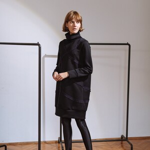 May include: A woman wearing a black turtleneck dress with a textured design and black tights. She is wearing black heels with a bow detail. The dress is knee-length and has a loose fit.