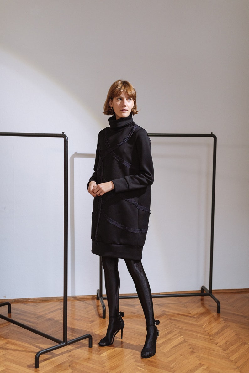 May include: A woman wearing a black turtleneck dress with a textured design and black tights. She is wearing black heels with a bow detail. The dress is knee-length and has a loose fit.