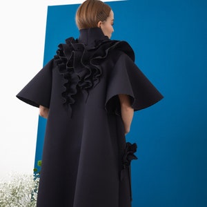 Pode incluir: Um casaco preto e fluido com gola alta e detalhes de babados. O casaco apresenta mangas largas e um elemento de design &uacute;nico de babados em cascata nas costas. A pe&ccedil;a de roupa est&aacute; sobre um fundo azul.
