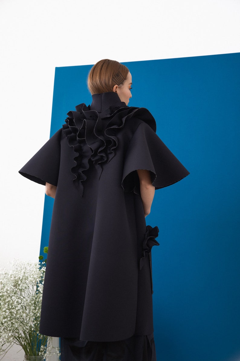 Pode incluir: Um casaco preto e fluido com gola alta e detalhes de babados. O casaco apresenta mangas largas e um elemento de design &uacute;nico de babados em cascata nas costas. A pe&ccedil;a de roupa est&aacute; sobre um fundo azul.