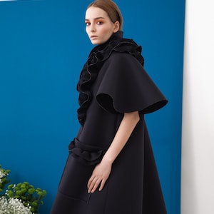 Pode incluir: Um casaco preto vanguardista com gola alta com babados e mangas amplas. O casaco apresenta um design &uacute;nico e em camadas com babados adicionais na cintura. O fundo &eacute; azul vibrante, criando um contraste marcante.