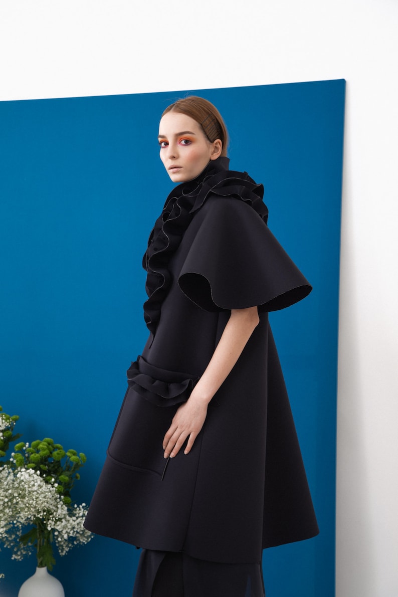 Pode incluir: Um casaco preto vanguardista com gola alta com babados e mangas amplas. O casaco apresenta um design &uacute;nico e em camadas com babados adicionais na cintura. O fundo &eacute; azul vibrante, criando um contraste marcante.