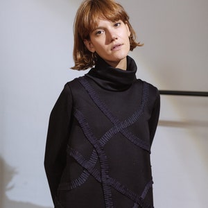 May include: A woman wearing a black turtleneck dress with intricate geometric designs created by ruched fabric.