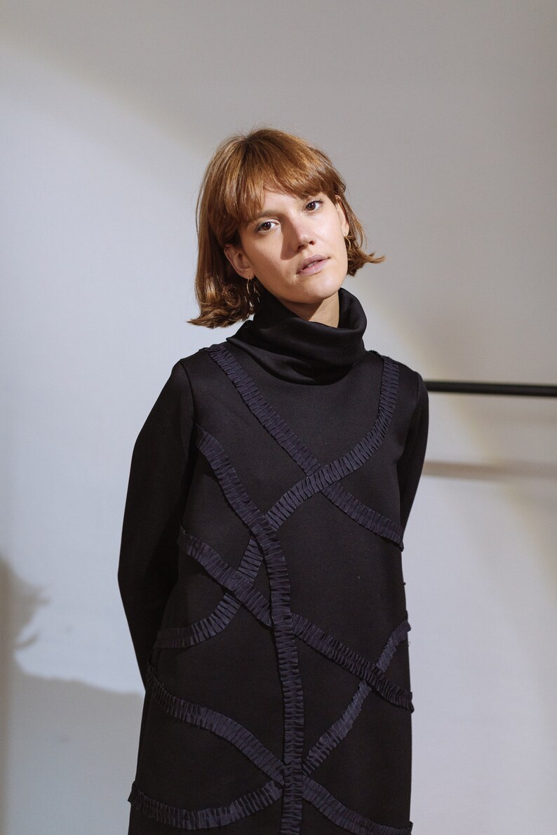 May include: A woman wearing a black turtleneck dress with intricate geometric designs created by ruched fabric.