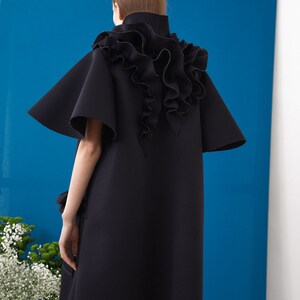 Pode incluir: Vestu&aacute;rio preto estilo capa com gola alta e mangas largas. As costas apresentam um grande detalhe com babados. A pe&ccedil;a est&aacute; sobre um fundo azul com detalhes brancos e um vaso de flores.