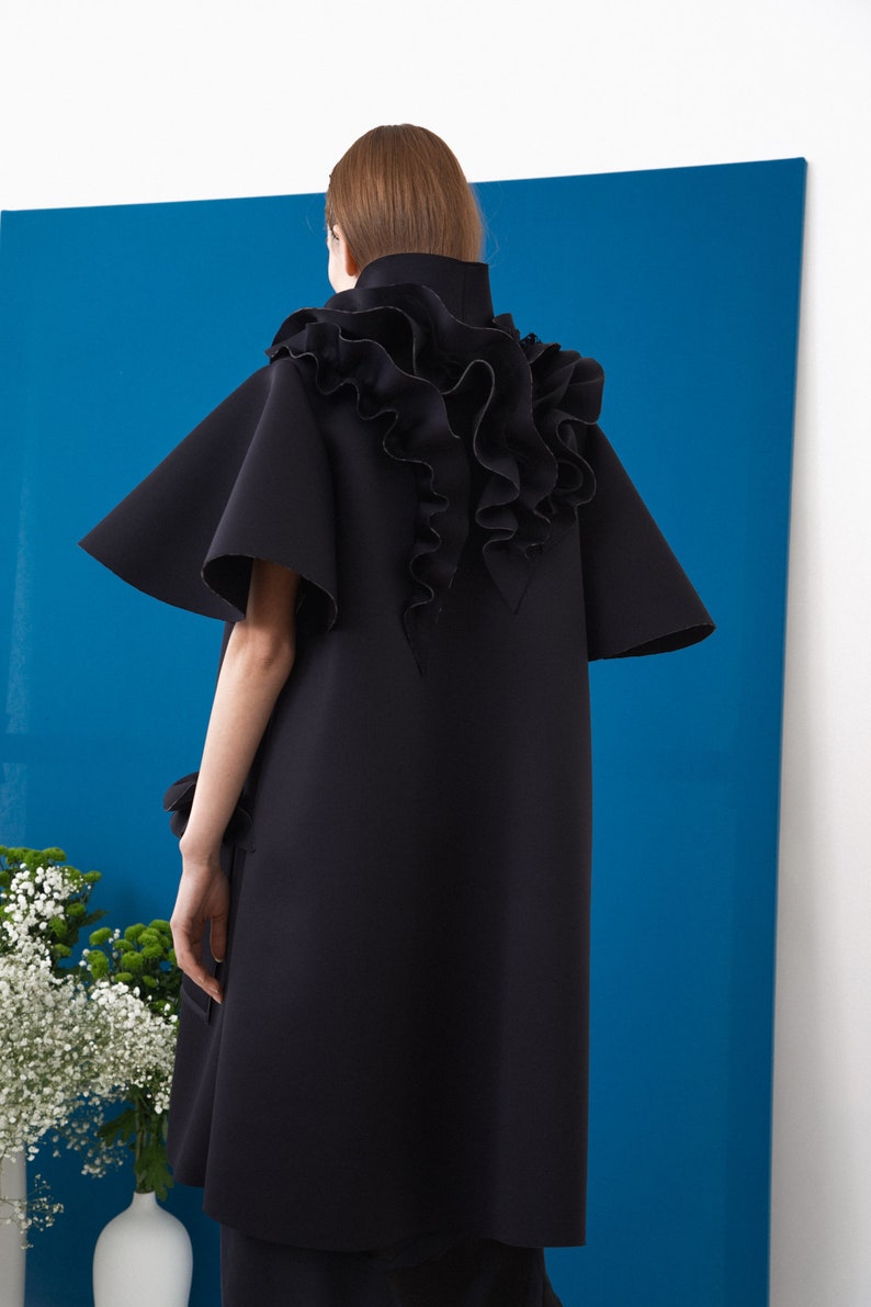 Pode incluir: Vestu&aacute;rio preto estilo capa com gola alta e mangas largas. As costas apresentam um grande detalhe com babados. A pe&ccedil;a est&aacute; sobre um fundo azul com detalhes brancos e um vaso de flores.