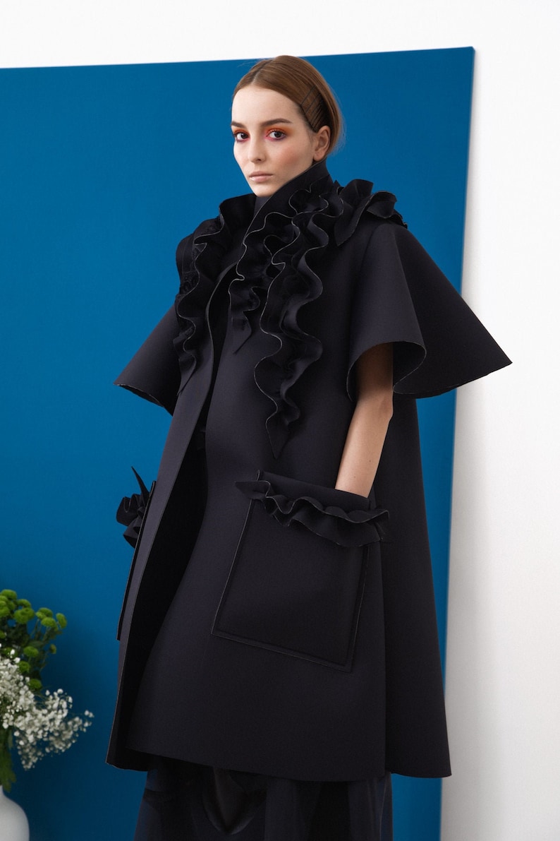 Pode incluir: Um casaco preto com um design &uacute;nico. O casaco apresenta uma gola alta com babados, mangas curtas e bolsos quadrados grandes com bordas com babados. O fundo &eacute; azul vibrante e branco.