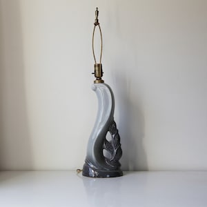 May include: A gray ceramic table lamp with a curved, stylized design. The lamp features a dark gray leaf-like pattern on the side and a brass-colored lamp holder. The lamp is approximately 18 inches tall.