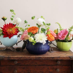 May include: Three vintage teacups filled with colorful flowers, including red, orange, pink, and white blooms. The teacups are displayed on a wooden table with drawers.
