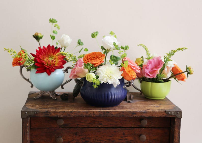 May include: Three vintage teacups filled with colorful flowers, including red, orange, pink, and white blooms. The teacups are displayed on a wooden table with drawers.