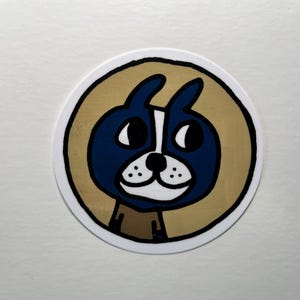 May include: A round sticker featuring a cartoon dog with a blue face, black eyes, and a white snout. The dog is set against a gold-colored circle with a black outline, on a white background. The dog has large, expressive eyes.
