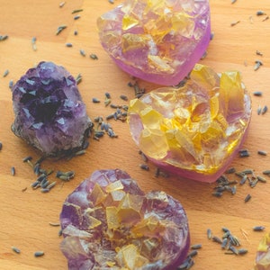 May include: Four heart-shaped soaps with a purple and gold crystal design. The soaps are on a wooden surface with dried lavender flowers scattered around them.