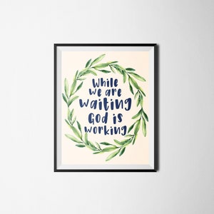 May include: A framed print with a cream background and a wreath of green leaves. The text "While we are waiting God is working" is written in blue inside the wreath.