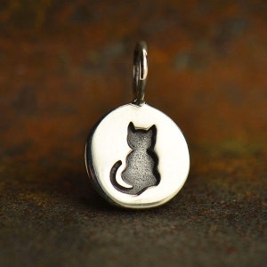 May include: A silver-colored pendant with a circular design featuring a cut-out silhouette of a cat. The cat is sitting with its tail curled. The pendant has a small loop at the top for attaching to a chain or bracelet. The background is a mottled brown.