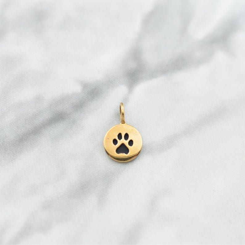 Puede incluir: Encanto de color dorado con un recorte de huella de pata negra. El encanto es de forma ovalada y tiene un bucle en la parte superior para sujetarlo a un collar o pulsera.