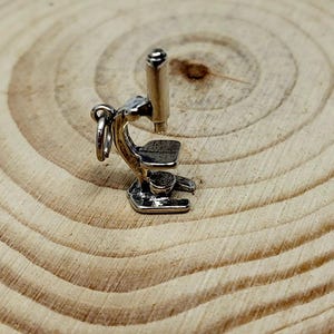 May include: A small, silver-colored microscope charm with a detailed design. The charm features a round eyepiece, a stage, and a base. The microscope is shown on a wooden surface.