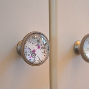 May include: Two silver cabinet knobs with clear glass inserts featuring a map design. The map is printed in black and white with pink accents. The knobs are attached to white cabinet doors.
