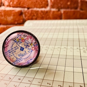 May include: A close-up of a small, round, glass paperweight with a map of Martha's Vineyard, Massachusetts. The map is printed in pink, blue, and black, and includes the numbers 26 and 336. The paperweight is sitting on a gridded notebook.