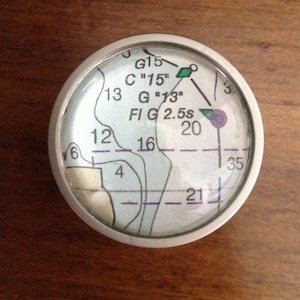 May include: A round, silver-colored compass with a white face and black markings. The compass face shows numbers 3, 6, 12, 13, 16, 20, 21, 35, and text that reads "G15", "C "15"", "G "13"", "FI G 2.5s".
