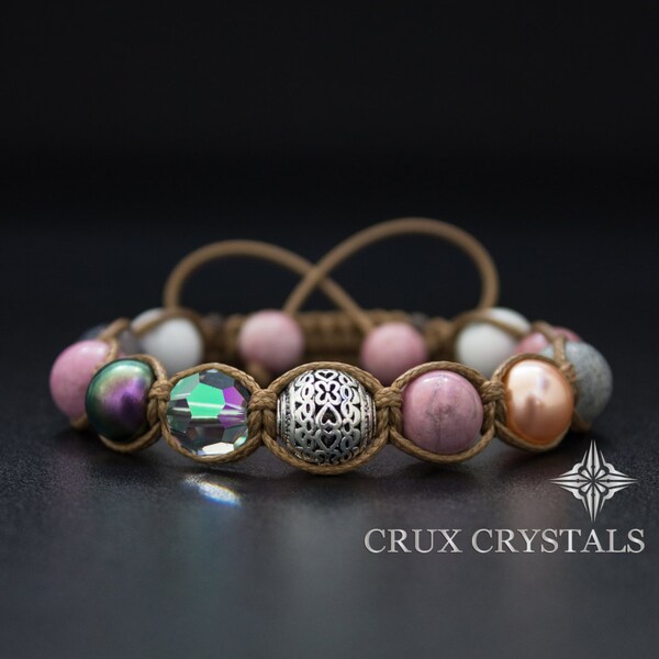 Op de afbeelding: Een bruin leren armband met roze, witte en grijze kralen, een zilveren kraal met een decoratief ontwerp en een heldere kristallen kraal. De armband ligt op een zwart oppervlak en de tekst "CRUX CRYSTALS" is zichtbaar onder de armband.