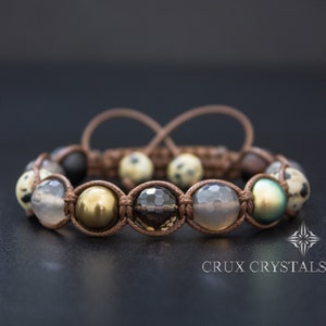 Op de afbeelding: Een bruin leren armband met een verscheidenheid aan edelsteen kralen, waaronder grijze agaat, bruin rookkwarts, goud en groene abalone. De armband is vastgemaakt met een bruin koord en heeft een knoopsluiting.