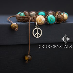May include: A brown leather cord bracelet with turquoise, brown, and white gemstone beads and a silver peace sign charm. The bracelet is tied with a knot and has a tassel on the end. The bracelet is on a black surface with the text "CRUX CRYSTALS" printed on it.