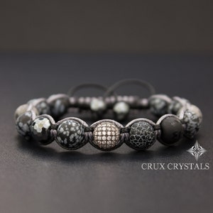 May include: A black and white beaded bracelet with a silver bead accented with clear cubic zirconia stones. The bracelet is made with a black cord and has a silver clasp. The bracelet is on a dark gray surface.  Crux Crystals.