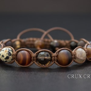 Op de afbeelding: Een bruin leren armband met verschillende edelsteen kralen, waaronder dalmatijnse jaspis, agaat en hematiet. De armband is met een knoop vastgemaakt en heeft een schuifsluiting.