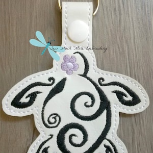 May include: White leather keychain with a black embroidered swirl design resembling a turtle with a purple flower on top. The keychain has a gold metal ring and a white button.