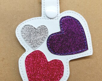Puede incluir: Llavero de cuero blanco con tres corazones de purpurina. Los corazones son plateados, morados y rosas.