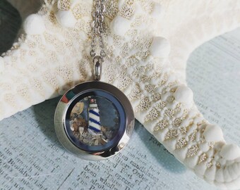 May include: Silver locket necklace with a lighthouse and sand inside. The lighthouse is blue and white with a red roof. The locket is open and the inside is visible.