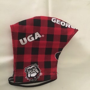 May include: A face mask with a red and black buffalo plaid pattern. The mask features the text "UGA GEORGIA" in white, along with a bulldog logo. The mask has black elastic ear loops.