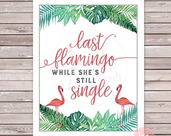 May include: A white sign with green tropical leaves around the border. The text on the sign reads "last flamingo while she's still single" in red cursive font. Two pink flamingos are on either side of the text.