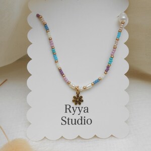 May include: A colourful beaded necklace with a gold flower pendant. The necklace has small beads in white, blue, purple, and gold. The tag reads "Ryya Studio".