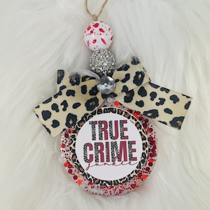 May include: A round, glittery ornament with the text "TRUE CRIME Junkie" in a white circle, accented with a leopard print bow. The ornament is topped with decorative beads and a jute string for hanging.