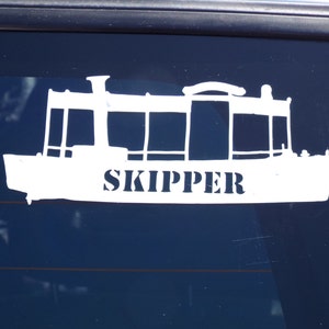 May include: White vinyl sticker of a boat with the word "SKIPPER" printed on the side. The sticker is on a dark blue car window.