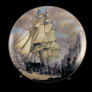 May include: Decorative plate featuring a detailed painting of a tall ship docked at a port. The artwork depicts the ship with billowing sails, figures on the dock, and buildings in the background. The plate has a gold rim.