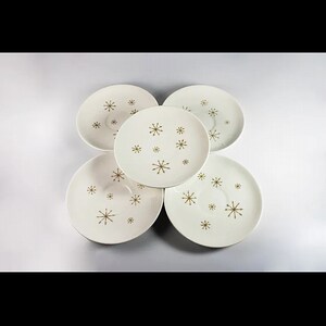 May include: Five white saucers with a gold starburst pattern. The saucers are arranged in a circular pattern, each displaying a different arrangement of the starburst design. The saucers are a vintage style.