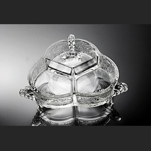 Puede incluir: Una fuente transparente para servir con tres secciones y tapa. La fuente tiene un diseño floral grabado y dos asas. La tapa tiene un asa decorativa. La fuente está sobre una superficie reflectante.