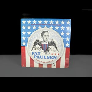 May include: Vintage vinyl record album cover featuring Pat Paulsen for President. The design incorporates a patriotic theme with a blue background of white stars, red and white stripes, and a central image of Paulsen.