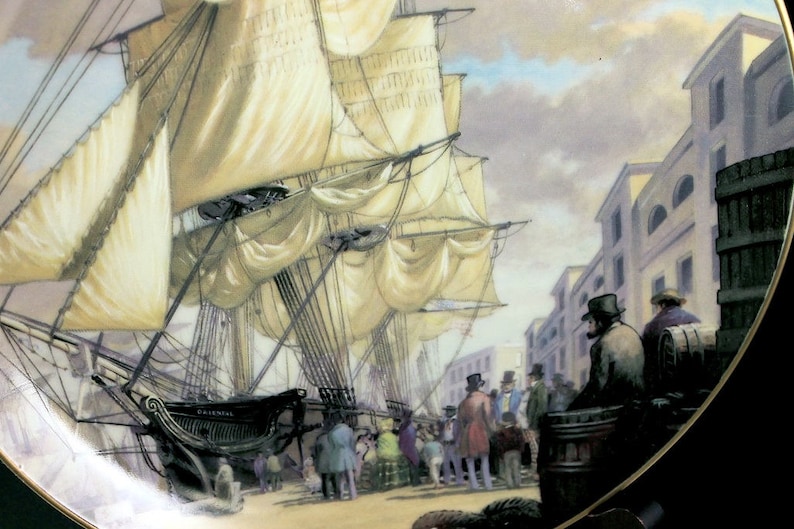 May include: Decorative plate featuring a detailed painting of a tall ship docked at a port. The ship has large white sails and is surrounded by people and buildings. The plate has a black border.