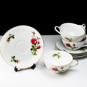 Peut inclure: Un ensemble de quatre tasses &agrave; th&eacute; et soucoupes blanches avec un motif floral repr&eacute;sentant des roses roses et des feuilles vertes. Les tasses et les soucoupes ont une bordure dor&eacute;e.