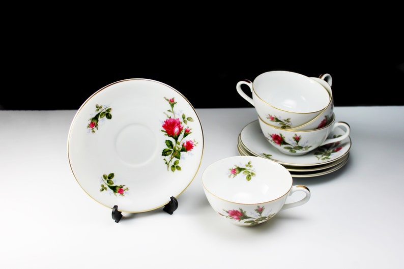 Peut inclure: Un ensemble de quatre tasses &agrave; th&eacute; et soucoupes blanches avec un motif floral repr&eacute;sentant des roses roses et des feuilles vertes. Les tasses et les soucoupes ont une bordure dor&eacute;e.